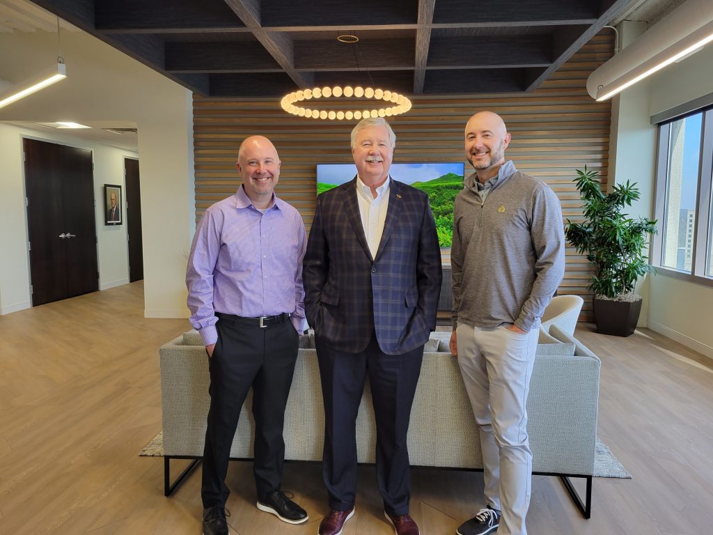 Andy, Jim and Patrick Roe standing in front of the new office. They are in front of a light grey couch. There is a TV in the background and a light above and behind them.