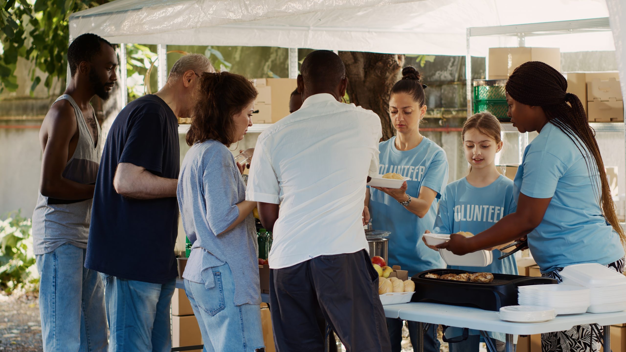 two women and one girl in blue shirts serving good to four adults, three men and one woman under a tent outside