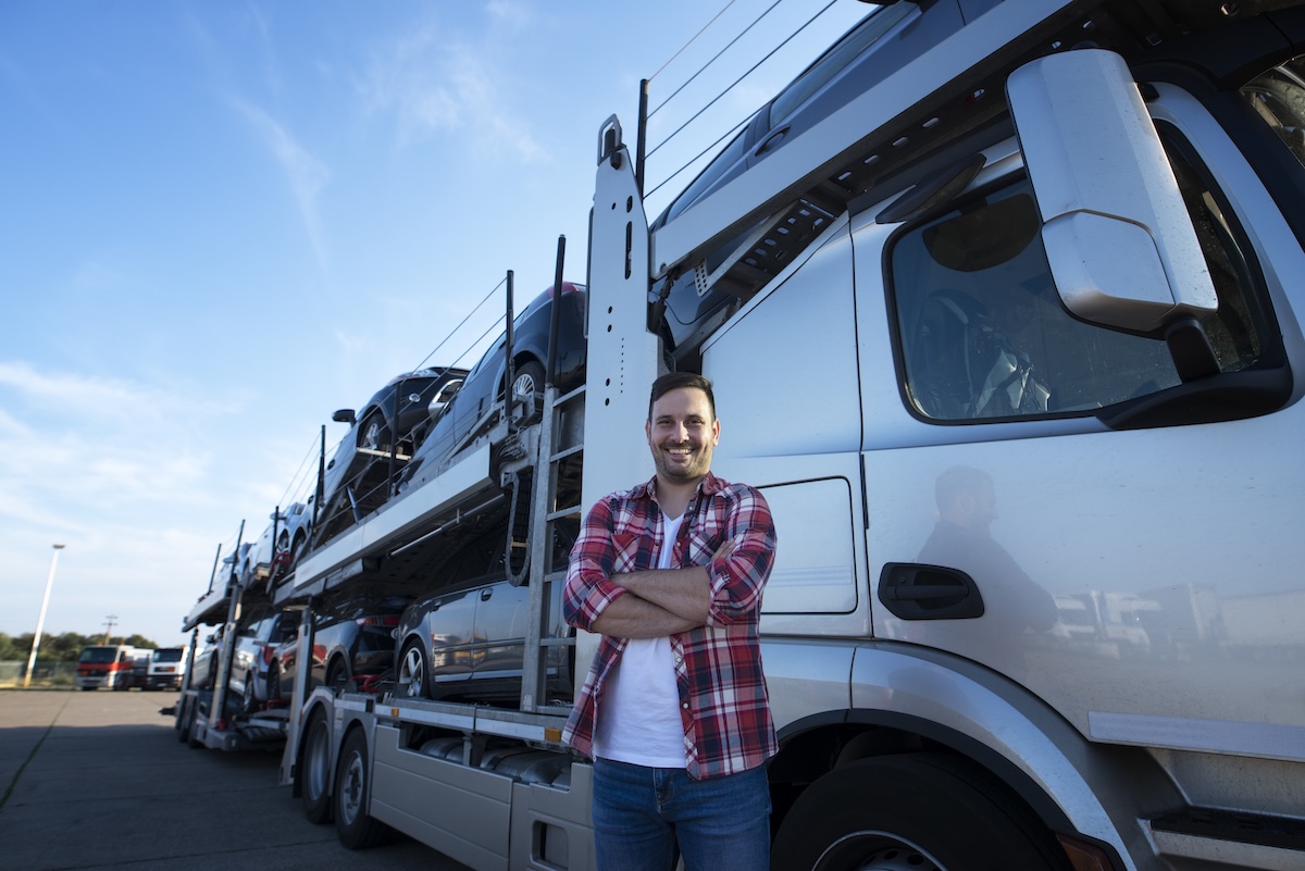 Portrait of professional smiling truck driver with crossed arms transporting cars to the market.