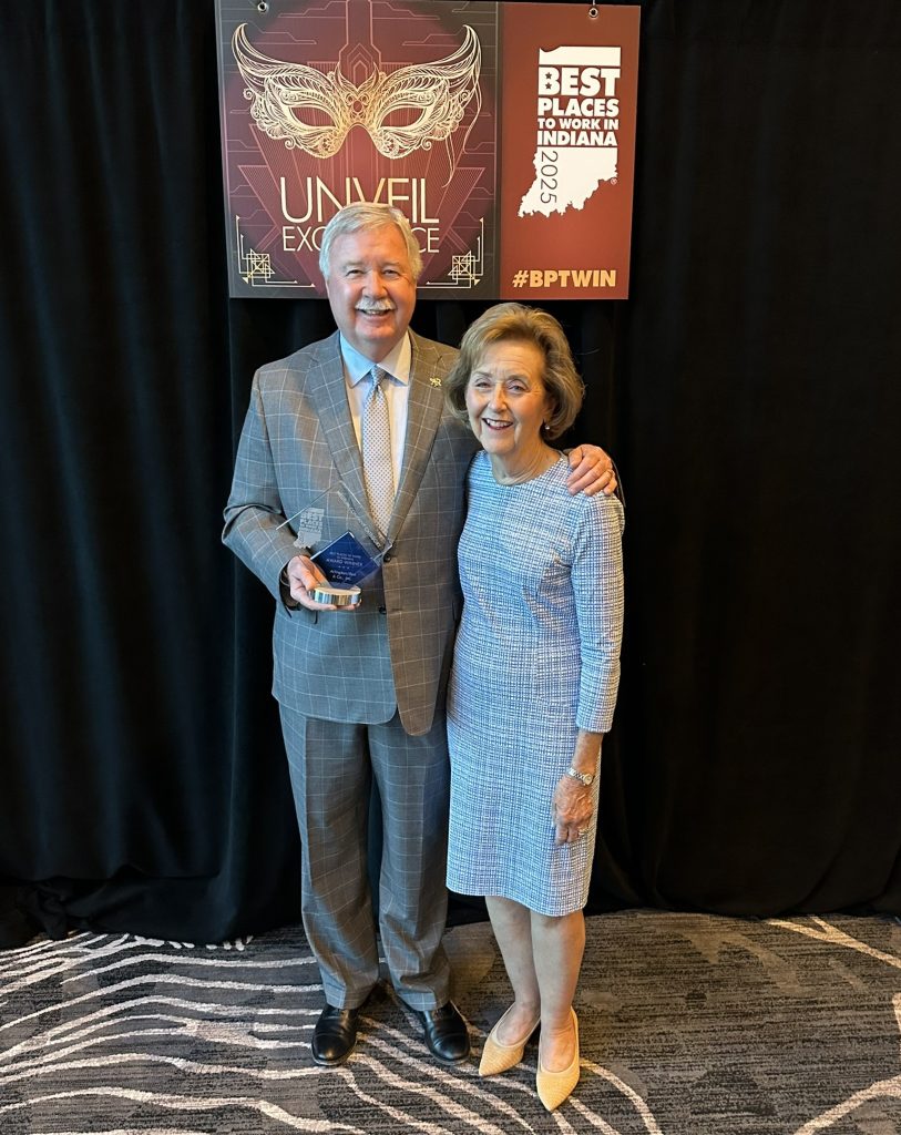 full body picture of jim roe and vickie roe smiling holding the best places to work in indiana 2025 award in front of black backdrop